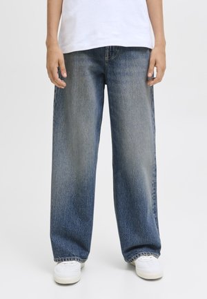 Person iført løstsiddende, falmede blå jeans, hvide sneakers og en hvid skjorte, der står mod en ensfarvet baggrund.