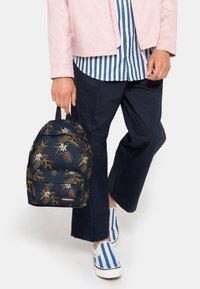 Marinblå ryggsäck med blommönster och bladmotiv, utrustad med en dragkedje-försedd framficka, vadderade axelremmar och vävd etikett. Strukturerad tyg.