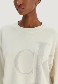 Pull crème clair avec une texture douce, un col côtelé et un logo gris bien visible sur le devant.