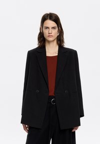 Zwarte dubbelknopen blazer van gestructureerde stof, met scherpe revers, twee knopen en een relaxed fit, gedragen over een roestkleurig top.