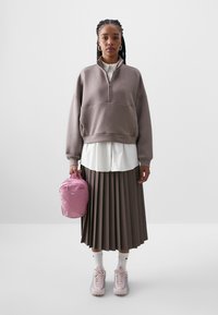 Túlméretezett barna cipzáras pulóver fehér inggel, pliszírozott barna szoknya, rózsaszín Nike hátizsák, fehér és rózsaszín sportcipők, valamint atlétazokni.