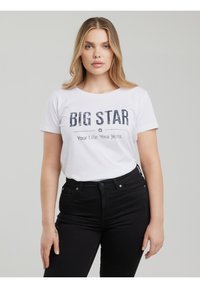 Blond naine, kes kannab valget T-särki, millel on kirjas "BIG STAR Your Life. Your Jeans.", ning kõrge vöökohaga musti teksaseid, seisab tavalise tausta ees.