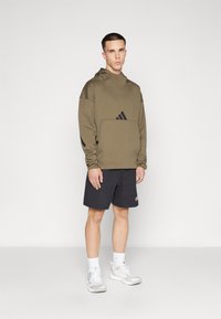 Felpa con cappuccio verde oliva con tasca a marsupio e dettagli in nero, abbinata a pantaloni corti neri e scarpe da ginnastica bianche.