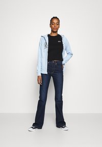 Světle modrá mikina na zip s logem, černé tričko, tmavě modré rozšířené džíny a černé tenisky s bílou podrážkou. Jednoduchý, každodenní outfit.