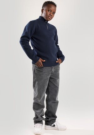 Marineblauwe gebreide trui met een ritssluiting bij de kraag, gecombineerd met grijze spijkerbroek en witte sneakers met zwarte accenten. Eenvoudig ontwerp, relaxte pasvorm.