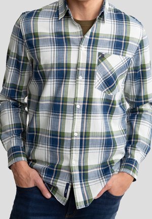 Homme portant une chemise à carreaux verte, bleue et blanche boutonnée avec une poche avant, mains dans les poches d'un jean bleu foncé, fond neutre.