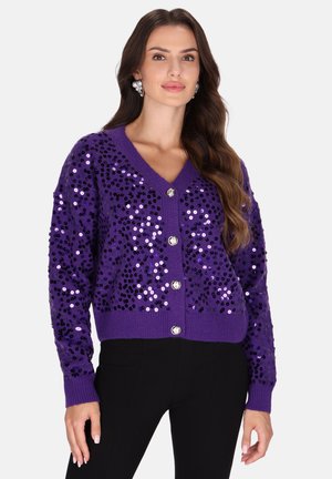Vrouw draagt een paarse cardigan met pailletten en knopen en zwarte broek, met lang golvend bruin haar en decoratieve oorbellen.