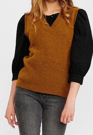 Femme portant un gilet sans manches tricoté couleur moutarde sur un chemisier noir à manches longues et un jean gris foncé, debout avec les bras détendus.