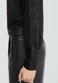 Czarna teksturowana bluzka z długim rękawem i bufkami, zestawiona z błyszczącymi czarnymi skórzanymi spodniami z zapięciem na guziki i boczną kieszenią.