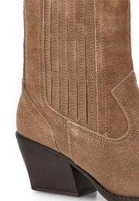 Botín de ante marrón hasta el tobillo con punta afilada y tacón apilado. Presenta costuras verticales y un diseño curvado en la costura.