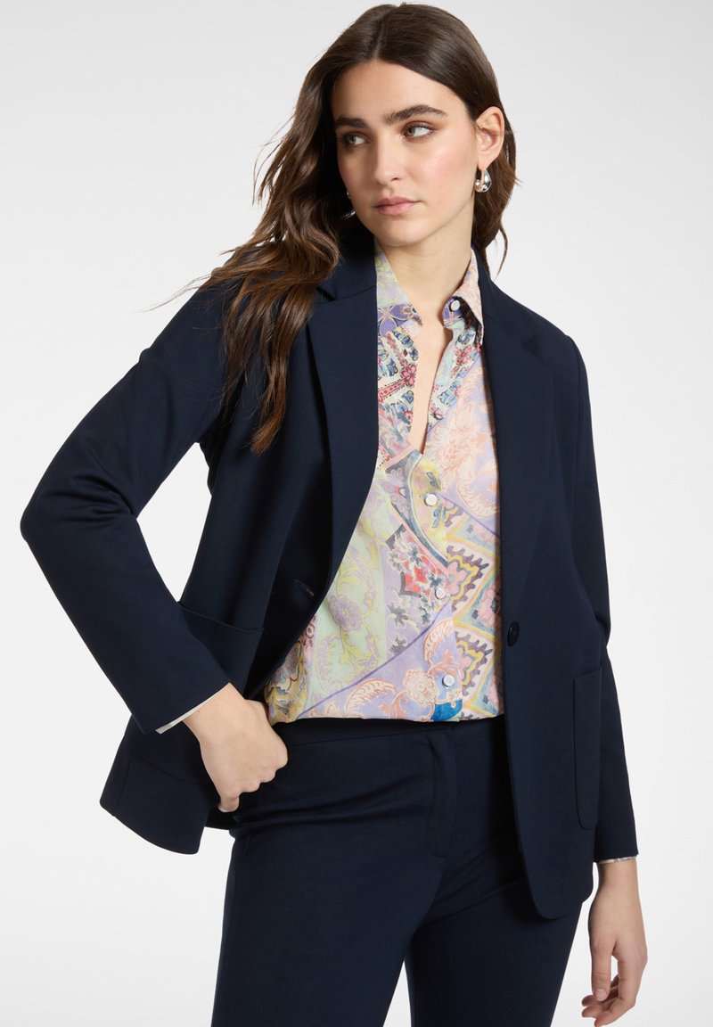 Mujer con cabello castaño ondulado, vestida con un blazer azul marino y pantalones, y una blusa de colores pastel con estampado, posando con una mano en el bolsillo.