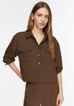 Kobieta o długich jasnobrązowych włosach nosi brązową koszulę zapinaną na guziki z dwoma kieszeniami na piersiach oraz pasujące brązowe spodnie, pozując na białym tle.