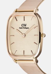 Kuldne ruudukujuline kell tekstureeritud kreemja diali, roosa tunni markerite ja võrksanga. Keskosas on brändi logo "Daniel Wellington".