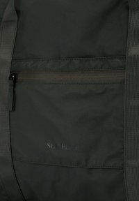 Bolsa de tela verde oscuro con un bolsillo frontal con cremallera, correas texturizadas y un logo sutil. El material es liso y duradero.