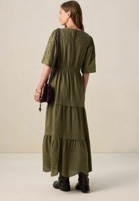 Robe à volants verte olive avec des manches courtes bouffantes, une taille élastique, des accents brodés et un détail en forme de trou de serrure au dos. Portée avec des bottines noires.
