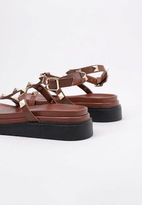Sandales en cuir marron avec des accents en pic doré, bride de cheville réglable et semelle épaisse en noir. Design à bout ouvert avec talon plat.