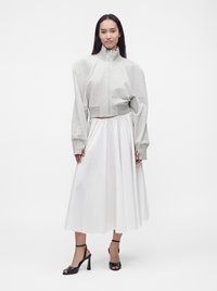 Femme portant une veste courte gris clair zippée, une jupe midi blanche et des sandales à talons noirs, debout sur un fond blanc.
