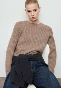 Brązowy sweter prążkowany z długim rękawem, wykonany z miękkiego materiału. Połączony z ciemnoniebieskimi dżinsami oraz ciemnym swetrem owiniętym w pasie.