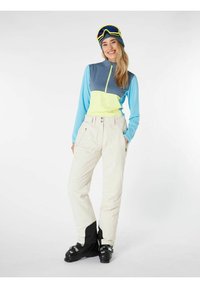 Skikleding bestaande uit een gekleurde, meerkleurige lange mouw top in blauw, grijs en geel, gecombineerd met witte skibroeken en zwarte skistokken.