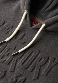 Sudadera gris con diseño texturizado en la parte frontal, cordones blancos y detalles metálicos. El material parece suave y tiene una forma clásica de sudadera.