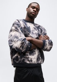 Νεαρός άνδρας με σταυρωμένα τα χέρια, φορώντας μαύρη και λευκή φανέλα με αφηρημένο μοτίβο και μαύρο παντελόνι, πάνω σε απλό φωτεινό φόντο.