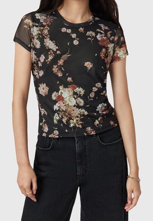 Frau trägt ein schwarzes transparentes kurzärmeliges Oberteil mit einem bunten Blumenmuster und hoch taillierte schwarze Jeans.