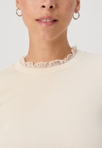 Ribbed, cream-colored top featuring a textured lace ruffle collar. Close-up view highlighting neckline and fabric details.