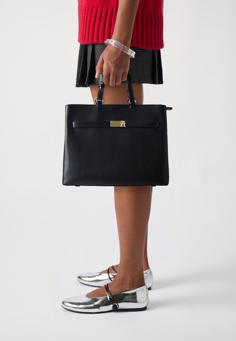 Personne en pull rouge et jupe noire tient un grand sac à main noir, porte des ballerines Mary Jane argentées et un bracelet transparent au poignet.