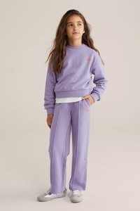 Svetlo lilac pulover in usklajene hlače iz mehkega blaga; vsebuje oranžni logotip na prsih in strani, sproščen kroj ter rebrasti manšeti. Srebrni športni copati.
