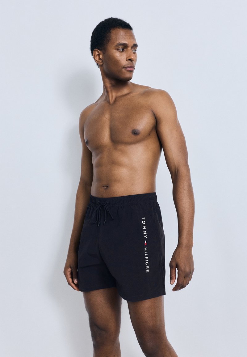 Μαύρα μαγιό σορτς με ελαστική μέση και κορδόνι, με λευκό λογότυπο στην αριστερή πλευρά. Λείο ύφασμα με κλασική εφαρμογή.