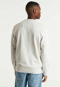Sudadera gris clara con cuello redondo y puños de canalé, que presenta un diseño limpio en la parte posterior y una textura suave. Llevada con jeans azules.
