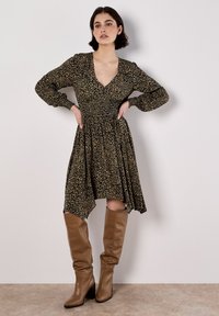 Schwarz-goldenes Leopardenmusterkleid mit langen Puffärmeln, einem tiefen V-Ausschnitt und einem gerafften Bund; kombiniert mit beigen bis zum Knie reichenden Stiefeln.