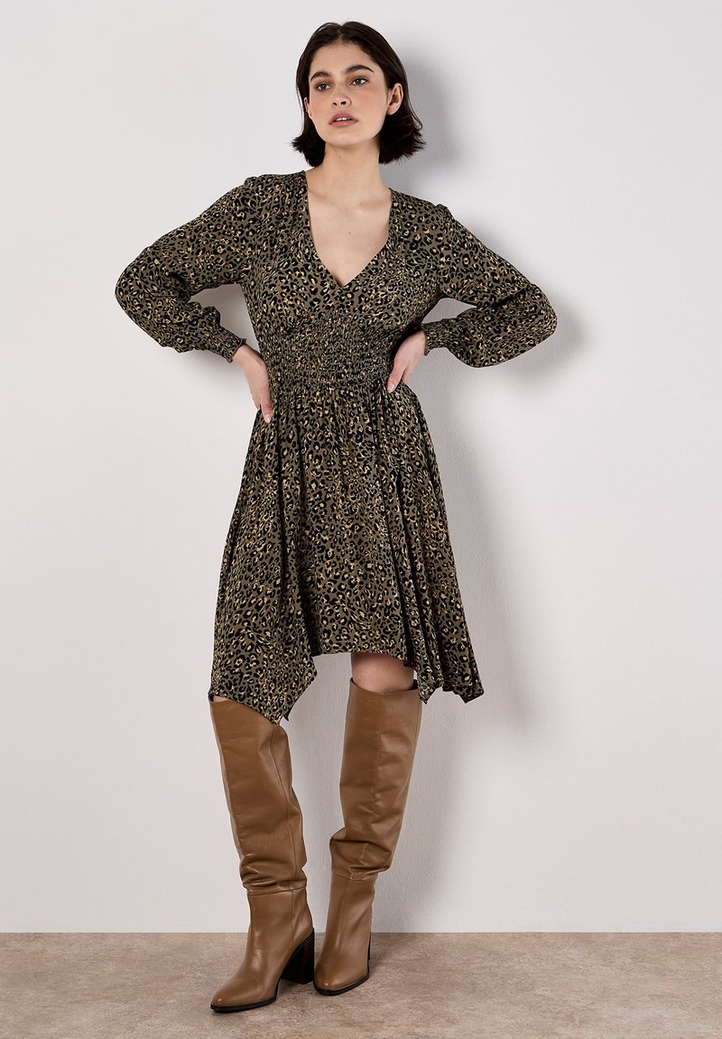 Schwarz-goldenes Leopardenmusterkleid mit langen Puffärmeln, einem tiefen V-Ausschnitt und einem gerafften Bund; kombiniert mit beigen bis zum Knie reichenden Stiefeln.