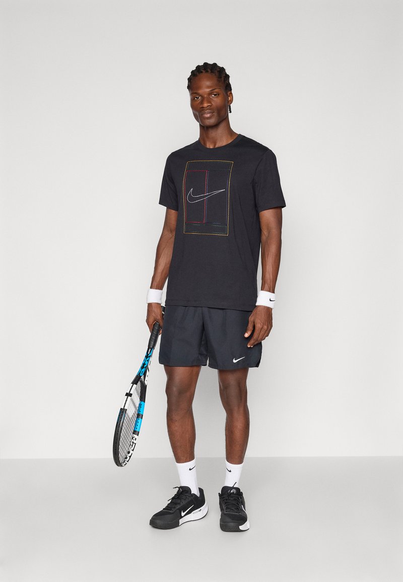 Zwart Nike t-shirt met een gekleurd rechthoekig logo-ontwerp, gecombineerd met zwarte shorts, witte sokken en zwarte Nike-schoenen, terwijl hij een tennisracket vasthoudt.