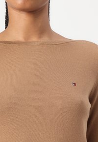 Maglione marrone a maglia con scollatura ampia e orli a coste, caratterizzato da una piccola etichetta logo in rosso, bianco e blu sul lato sinistro del petto.