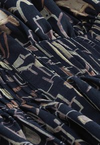 Textilní oděv s tmavým pozadím, představující smíšený abstraktní vzor v hnědé a béžové. Texturovaný, měkký materiál s nařasenými designovými detaily.