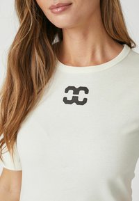 Light beige t-shirt with a round neckline, short sleeves, and a black abstract logo printed on the front. Soft, smooth cotton fabric.
