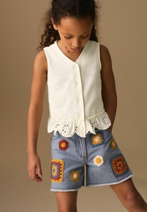 Mladé dievča nosí biely bezrukávový top na gombíky s očkovými volánmi a jeansové šortky zdobené farebnými háčkovanými kvetinovými záplatami.