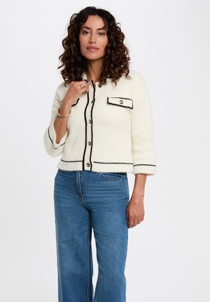 Cardigan accorciato color crema realizzato in materiale morbido con finiture nere, dotato di una tasca frontale, bottoni rotondi dorati e maniche a tre quarti.