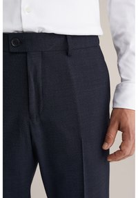 Uomo che indossa pantaloni da uomo blu scuro con trama, pince anteriori e bottone nero, abbinati a una camicia bianca a maniche lunghe.