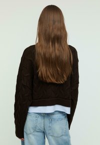 Maglione marrone lavorato a maglia con motivo a trecce, design cropped e maniche ampie, indossato sopra una camicia azzurro chiaro e abbinato a jeans chiari.