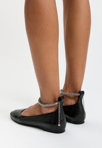Bailarinas de cuero negro con punta afilada y una cadena de acento en la parte posterior adornada con pequeños rhinestones, presentando una suela baja y plana.