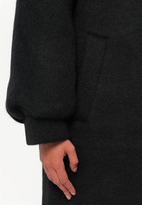 Cappotto di lana nera con una texture morbida, maniche lunghe a sbuffo e tasche laterali, con una mano appoggiata sul tessuto per il confronto delle dimensioni.