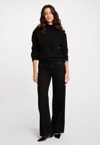 Suéter negro de punto con cuello alto, que presenta un acabado texturizado y botones decorativos en un hombro, combinado con pantalones anchos negros.