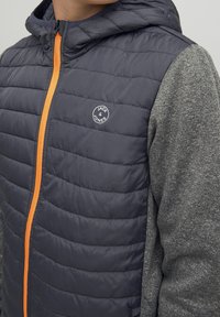 Kurtka z kapturem o pikowanej granatowej korpusie, szarych rękawach i pomarańczowych detalach zamka. Na lewej piersi znajduje się okrągłe szare logo.
