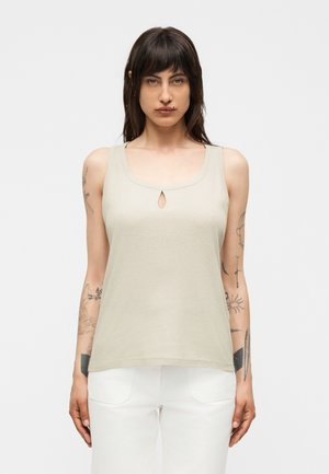 Sötét hajú felnőtt, aki bézs színű, ujjatlan, kulcscsontos részlettel ellátott felsőt és fehér nadrágot visel, egyszínű fehér háttér előtt áll.