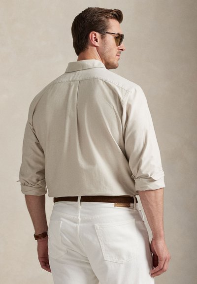 Beige skjorte med krage og oppbrettede ermer, kombinert med hvite bukser. Viser bakfra med brun belte og synlige lommer.