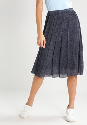 Vrouw draagt een marineblauwe geplooide midi-rok met kleine stippenpatronen en witte slip-on sneakers, staand tegen een effen achtergrond.