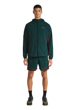 Mandlig model iført mørkegrøn let hættejakke og matchende shorts med sorte og olivengrønne sneakers på hvid baggrund.