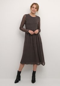 Långärmad klänning, mörk bakgrund med geometriskt mönster i brunt och beige. Halvlång, flödande tyg. Modell som bär ankelstövlar.